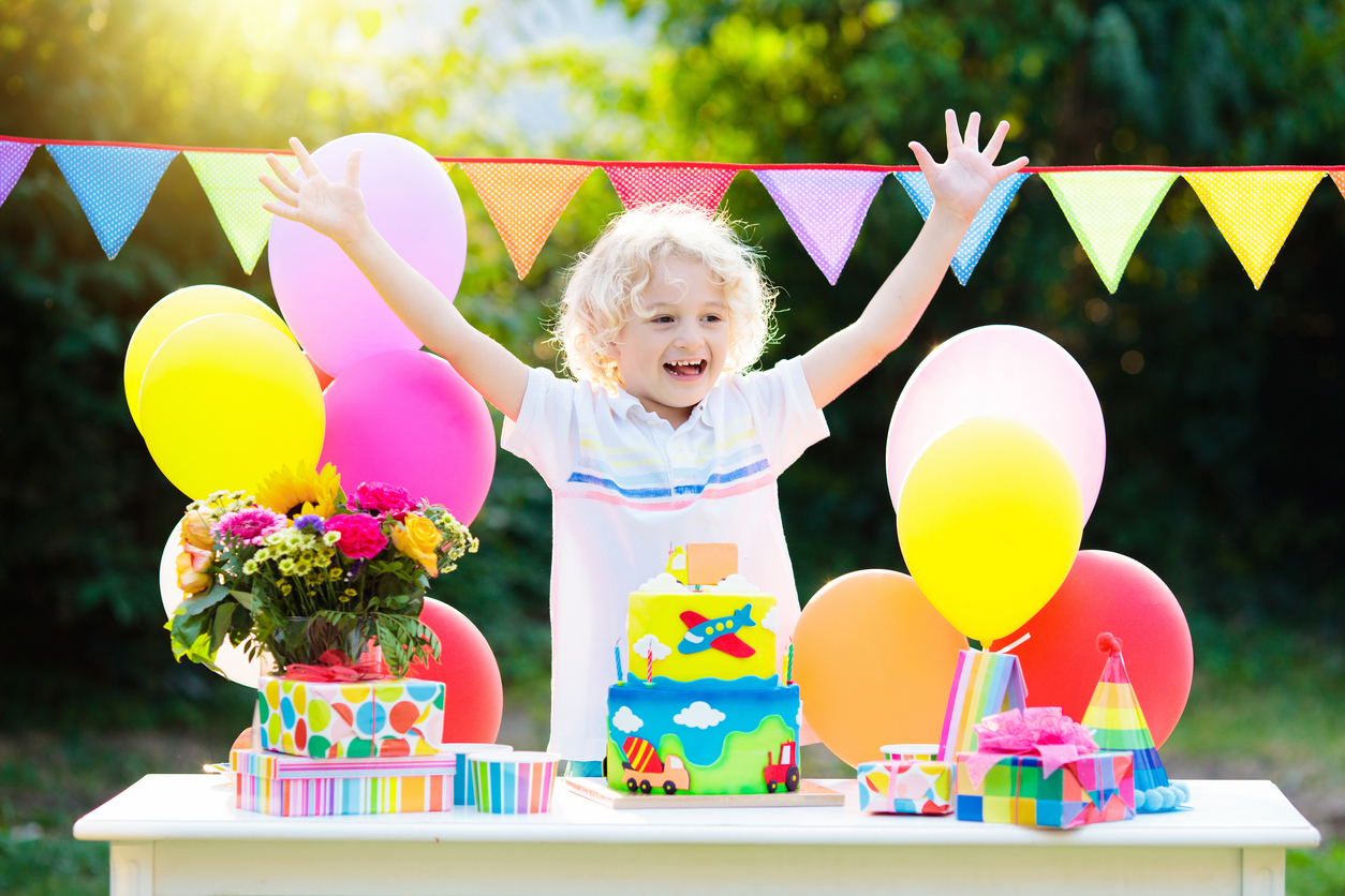 Kids birthday party. Child blowing out candles on colorful cake. Decorated garden with rainbow flag banners, balloons. Car and airplane theme celebration. Little boy celebrating birthday. Party food.