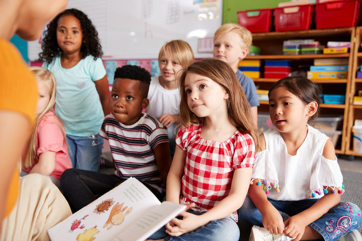Group Of Elementary School Pupils Sitting On Floor Listening To Female Teacher Read Story Group Of Elementary School Pupils Sitting On Floor Listening To Female Teacher Read Story