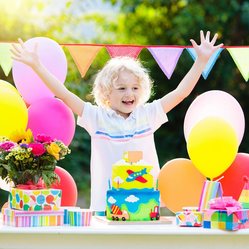 Kids birthday party. Child blowing out cake candle Kids birthday party. Child blowing out candles on colorful cake. Decorated garden with rainbow flag banners, balloons. Car and airplane theme celebration. Little boy celebrating birthday. Party food.