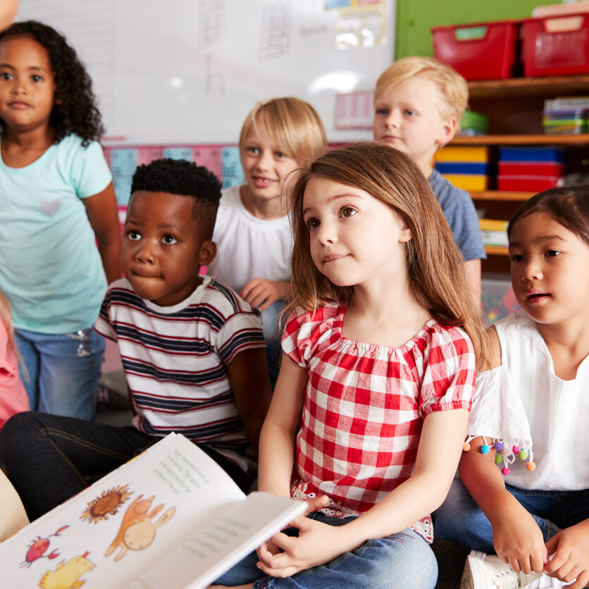 Group Of Elementary School Pupils Sitting On Floor Listening To Female Teacher Read Story Group Of Elementary School Pupils Sitting On Floor Listening To Female Teacher Read Story